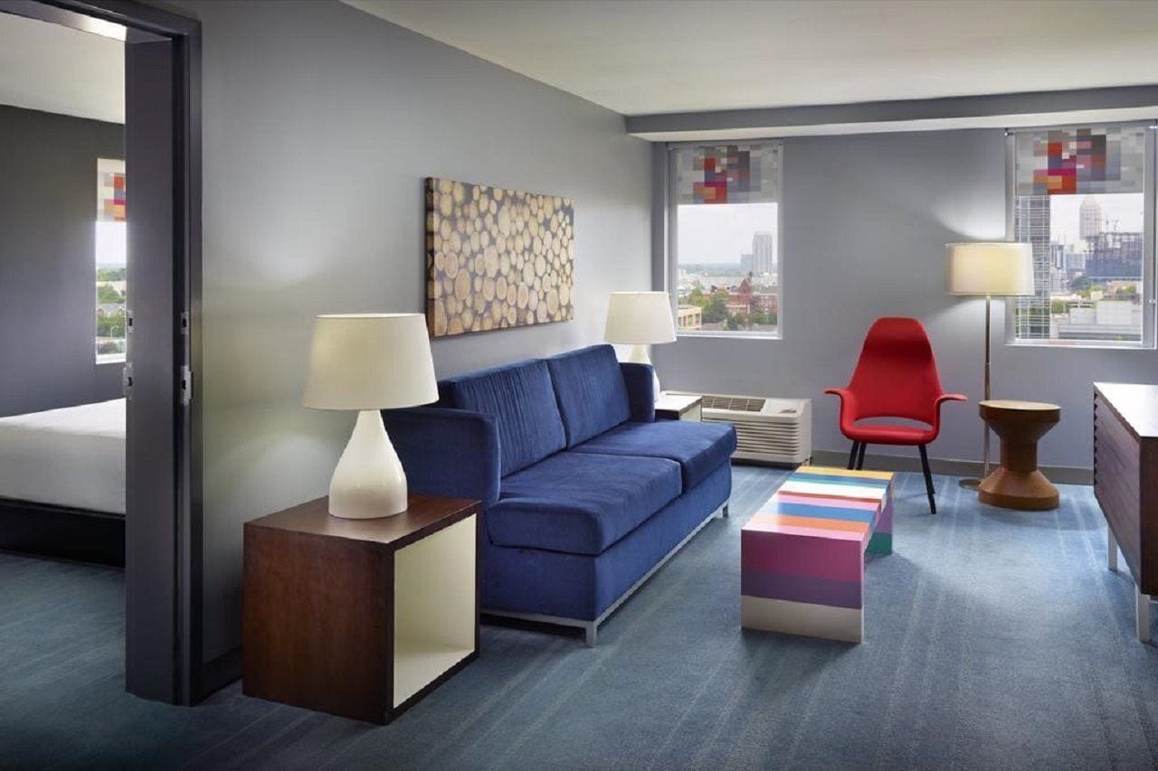 A modern hotel room with a blue sofa, two bedside lamps, a colorful ottoman, a red chair, and a city view through large windows.