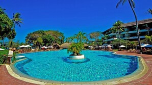 Une grande piscine aux eaux cristallines, entourée de palmiers et de chaises longues, avec un bâtiment visible au loin.
