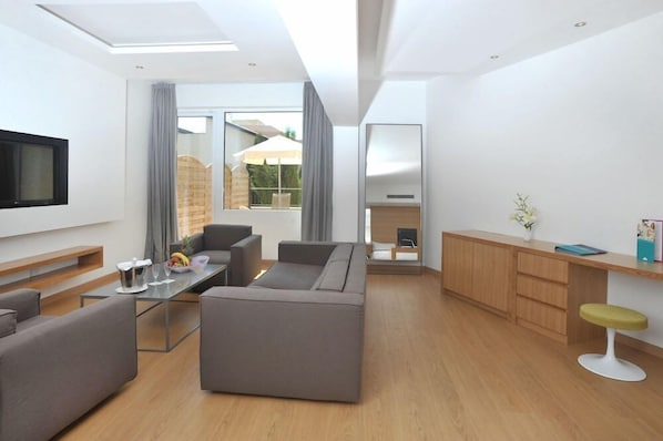 A modern living room with a sofa, a coffee table, and a TV. There is a window with curtains, a wooden cabinet, and a small stool. - Napa Plaza Hotel-adults Only (AYIA NAPA)