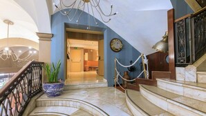 A grand staircase with marble steps leading to a landing, a blue wall with a clock, and a chandelier. - Del Mar Hotel (Barcelona)