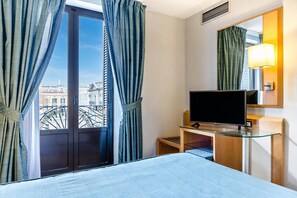 A hotel room with a bed, a TV on a glass table, a window with curtains, and a view of buildings. - Del Mar Hotel (Barcelona)