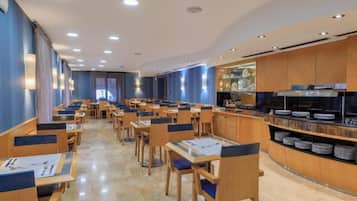 Un restaurante moderno con mesas y sillas largas, una zona de bufé con platos, y una cocina al fondo.