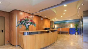 A reception area with a wooden reception desk, a large floral arrangement, a seating area, and a mural on the wall. - Del Mar Hotel (Barcelona)