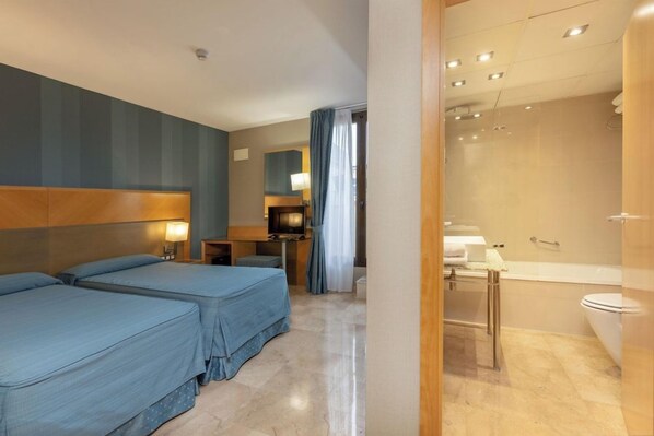 A hotel room with two beds, a desk, a TV, and a bathroom with a bathtub and toilet. - Del Mar Hotel (Barcelona)
