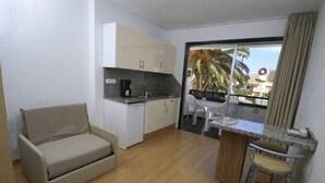 A compact living space with a kitchenette, a sofa, a dining area with a bar stool, and a balcony with a view of palm trees and buildings. - Koka Apartamentos (San Bartolomé de Tirajana)