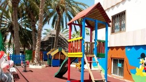 A playground with colorful play structures, palm trees, and a mural of cartoon characters on a building. - Koka Apartamentos (San Bartolomé de Tirajana)