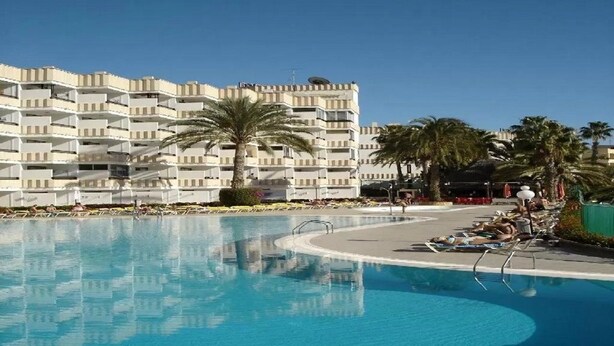 A swimming pool with lounge chairs and palm trees, surrounded by a multi-story building with balconies. - Koka Apartamentos (San Bartolomé de Tirajana)