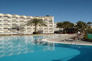 A swimming pool with lounge chairs and palm trees, surrounded by a multi-story building with balconies. - Koka Apartamentos (San Bartolomé de Tirajana)