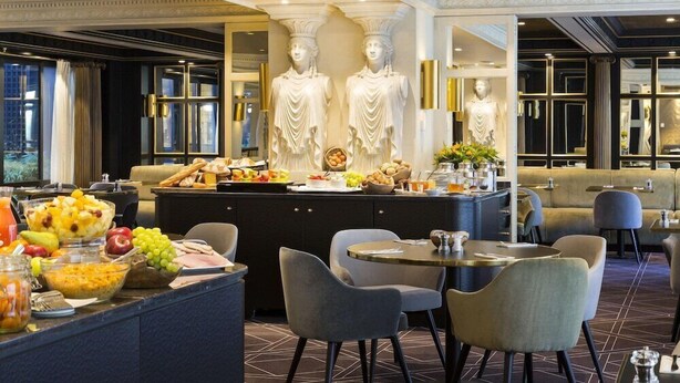 Un hall d’hôtel doté d’un bar central, de différents espaces aménagés pour s’asseoir et de statues classiques.