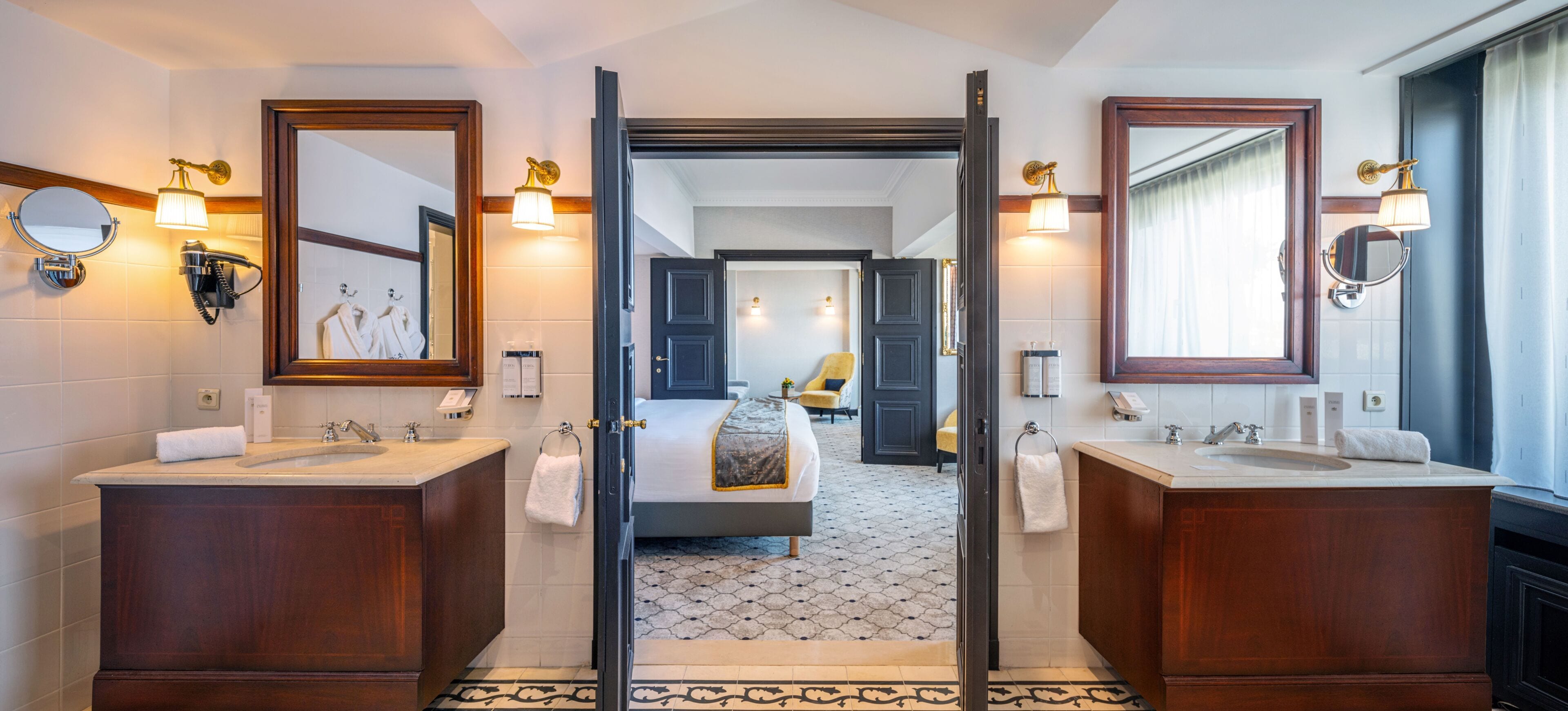 Een hotelbadkamer met twee wasbakken, een spiegel, een handdoekhouder en een deur naar een slaapkamer.