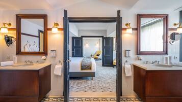 Een hotelbadkamer met twee wasbakken, een spiegel, een handdoekhouder en een deur naar een slaapkamer.