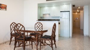 Un comedor con una mesa redonda y seis sillas, una cocina con armarios blancos y un refrigerador de acero inoxidable, y un pasillo que conduce a una puerta.