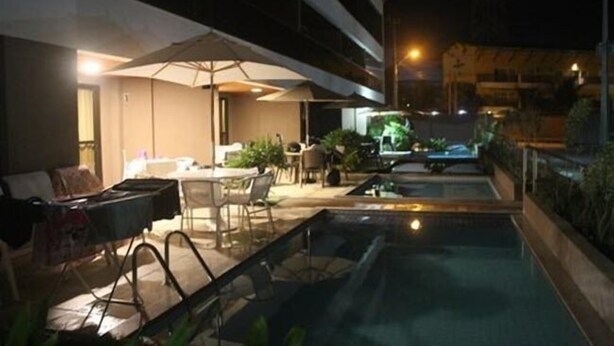 An outdoor pool area with lounge chairs and umbrellas, surrounded by greenery and a building in the background. - CPG02 Flat Piscina Privada em Porto (Ipojuca)