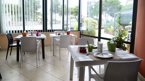 A dining area with white tables and chairs, large windows with blinds, and potted plants. - B & B Paradise (Melendugno)