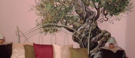 A bedroom with a bed, pillows, a nightstand, and a wall mural of a tree.
