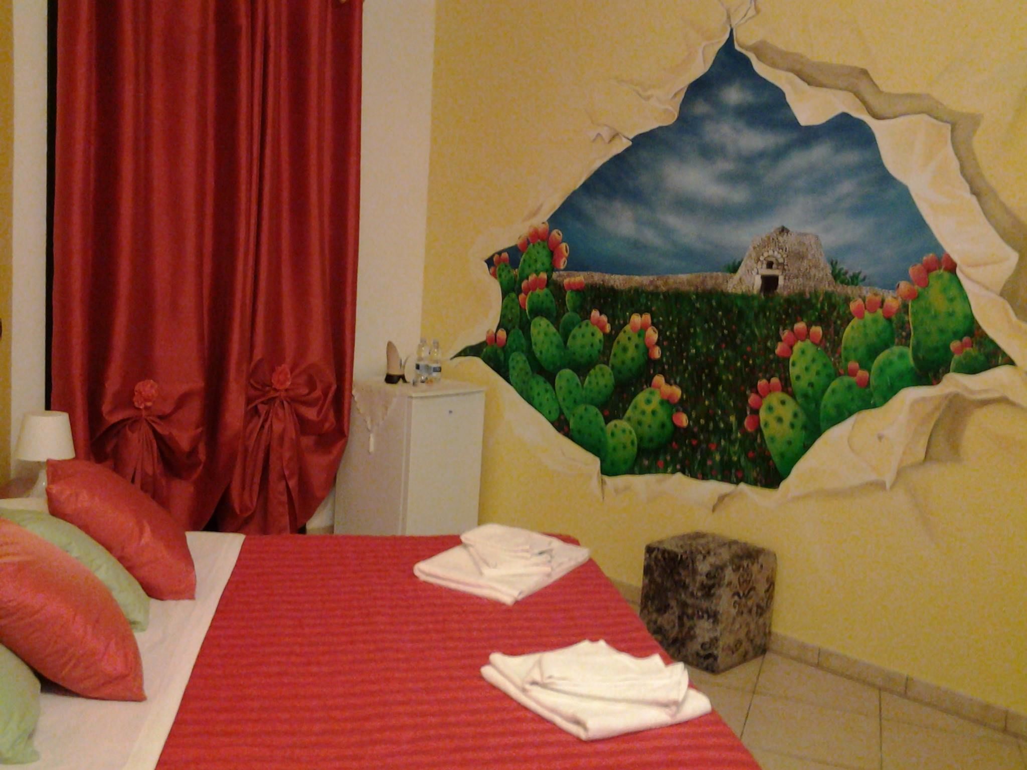 A hotel room with a bed, red curtains, a wall mural of a landscape with cacti and a small building, and a white cabinet.