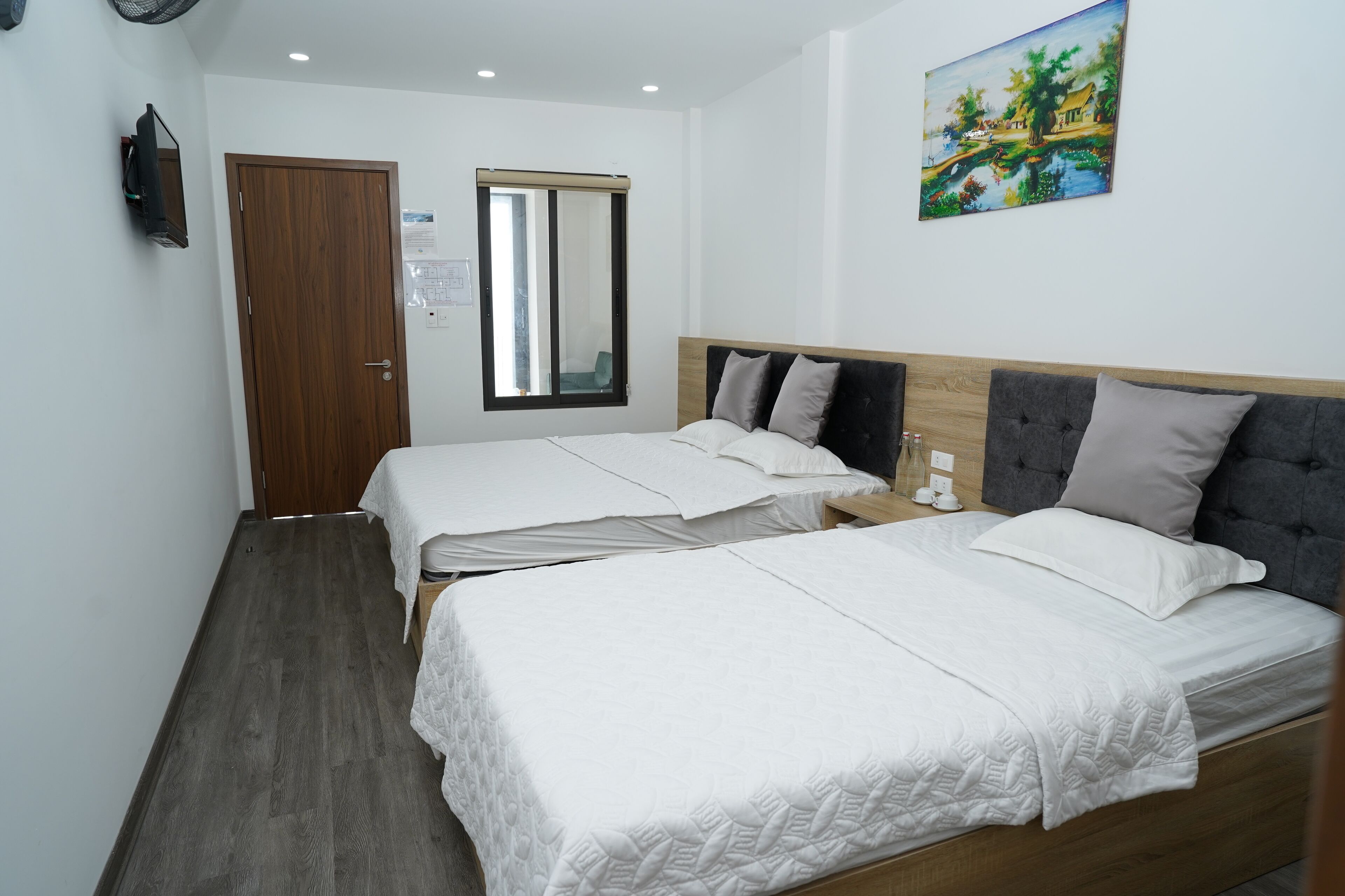 Une chambre d’hôtel avec deux lits, une porte en bois, une fenêtre avec des stores, un tableau au mur et une petite table avec une lampe.