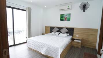 A modern hotel room with a large bed, a wooden headboard, a bedside table, a nightstand, a wall-mounted air conditioner, a ceiling fan, and a large window with a view of the outside.