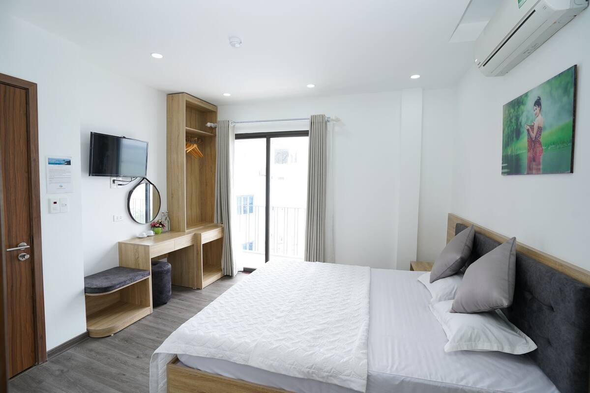 A modern hotel room with a large bed, a TV mounted on the wall, a wooden wardrobe, and a balcony with curtains.