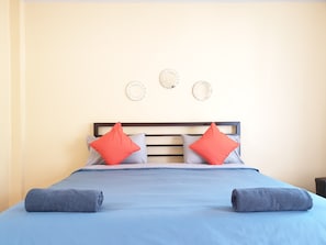 A neatly made bed with a blue bedspread, two pillows (one red, one grey), and two rolled towels.