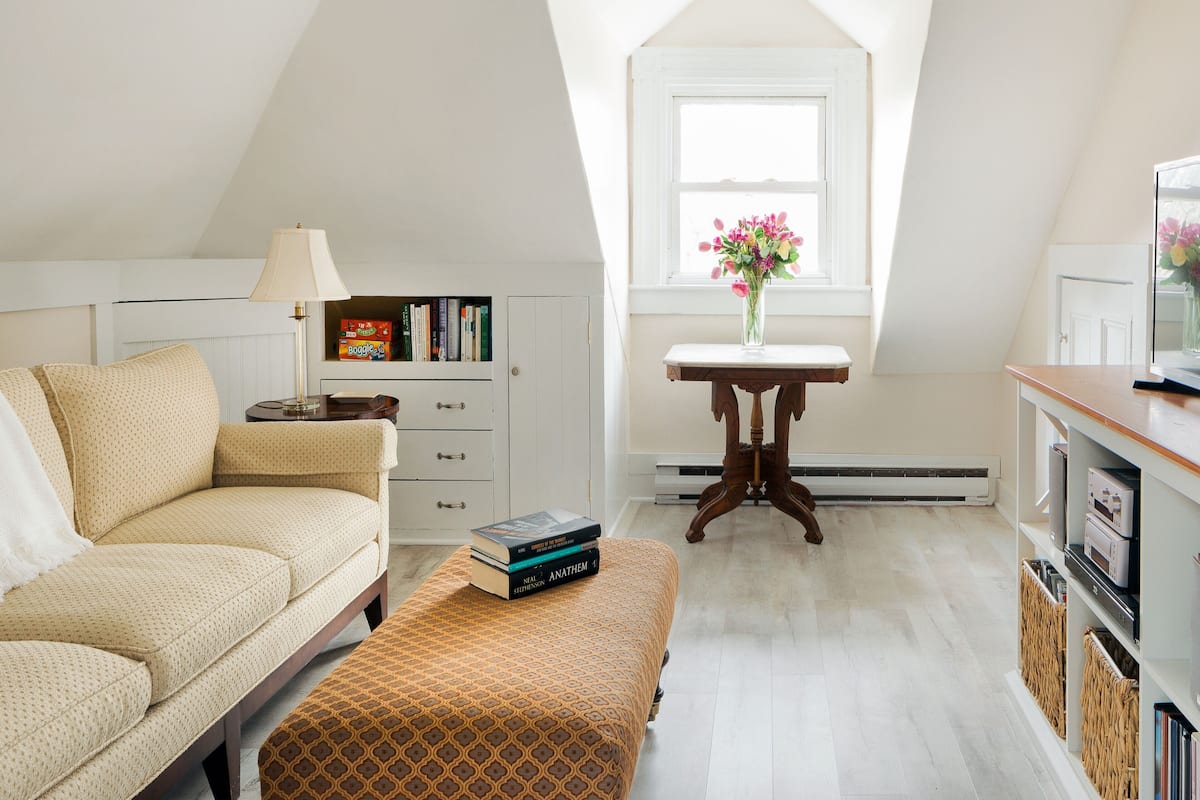 A cozy attic bedroom with a sloped ceiling, a sofa, a small table with books, a window with flowers, and a TV on a wooden stand.