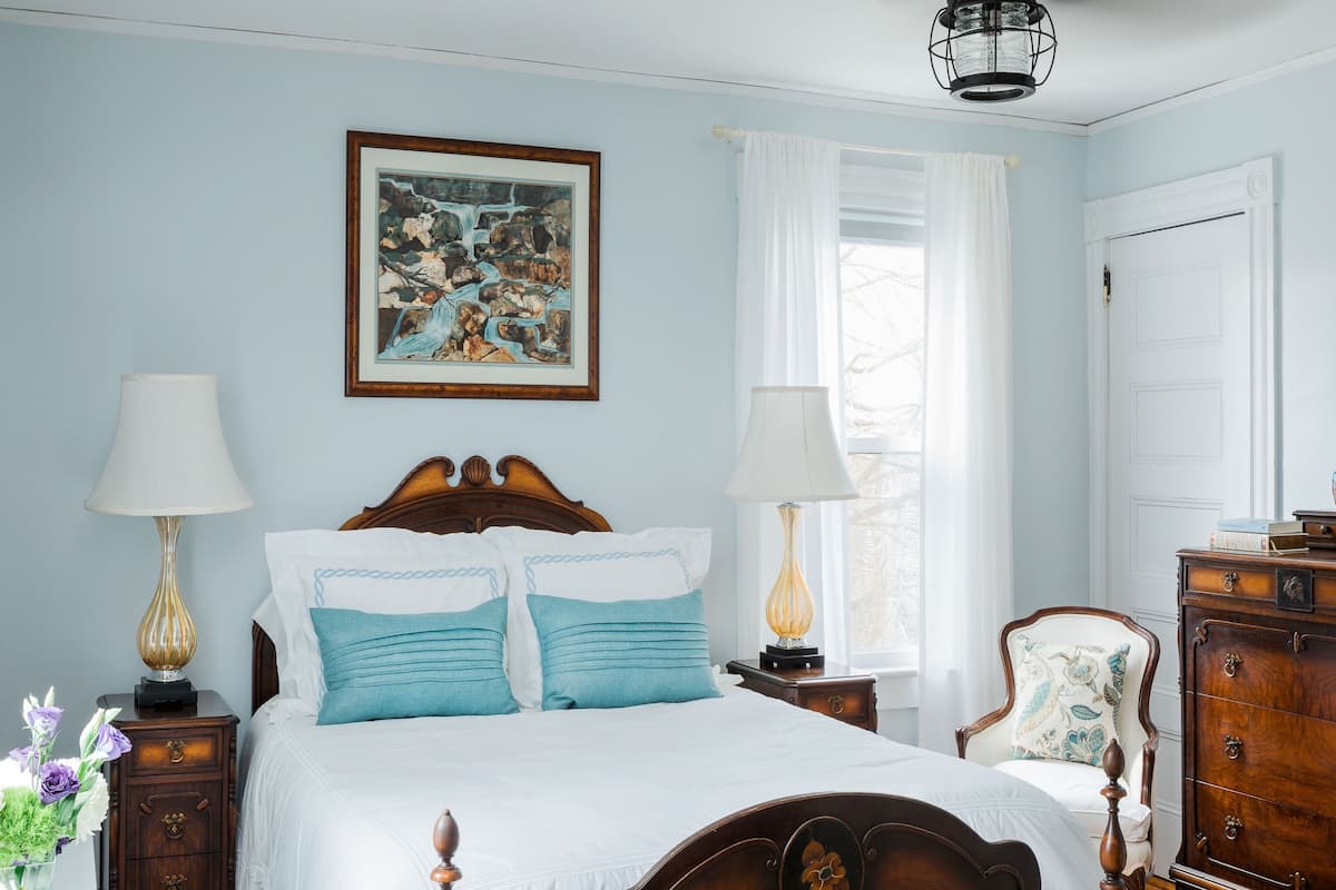 A bedroom with a wooden bed, two bedside tables with lamps, a nightstand with a vase of flowers, and a window with curtains.