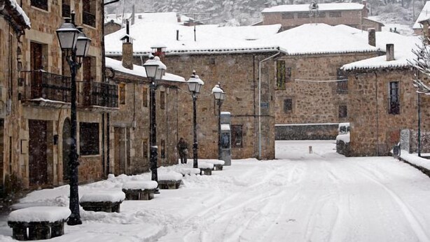Una calle nevada con edificios de piedra, bancos y farolas.