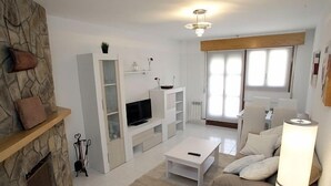 Una sala de estar moderna con chimenea de piedra, muebles blancos, un televisor de pantalla plana y un ventanal con persianas.