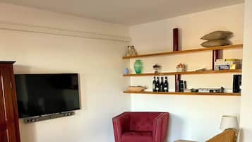 Ein Wohnzimmer mit einem Flachbildfernseher, einem roten Sessel, einem beigen Sofa mit gemustertem Überwurf und einem Couchtisch.