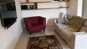 Una sala de estar con un sillón rojo, un sofá color beige, una alfombra estampada y un televisor montado en la pared.