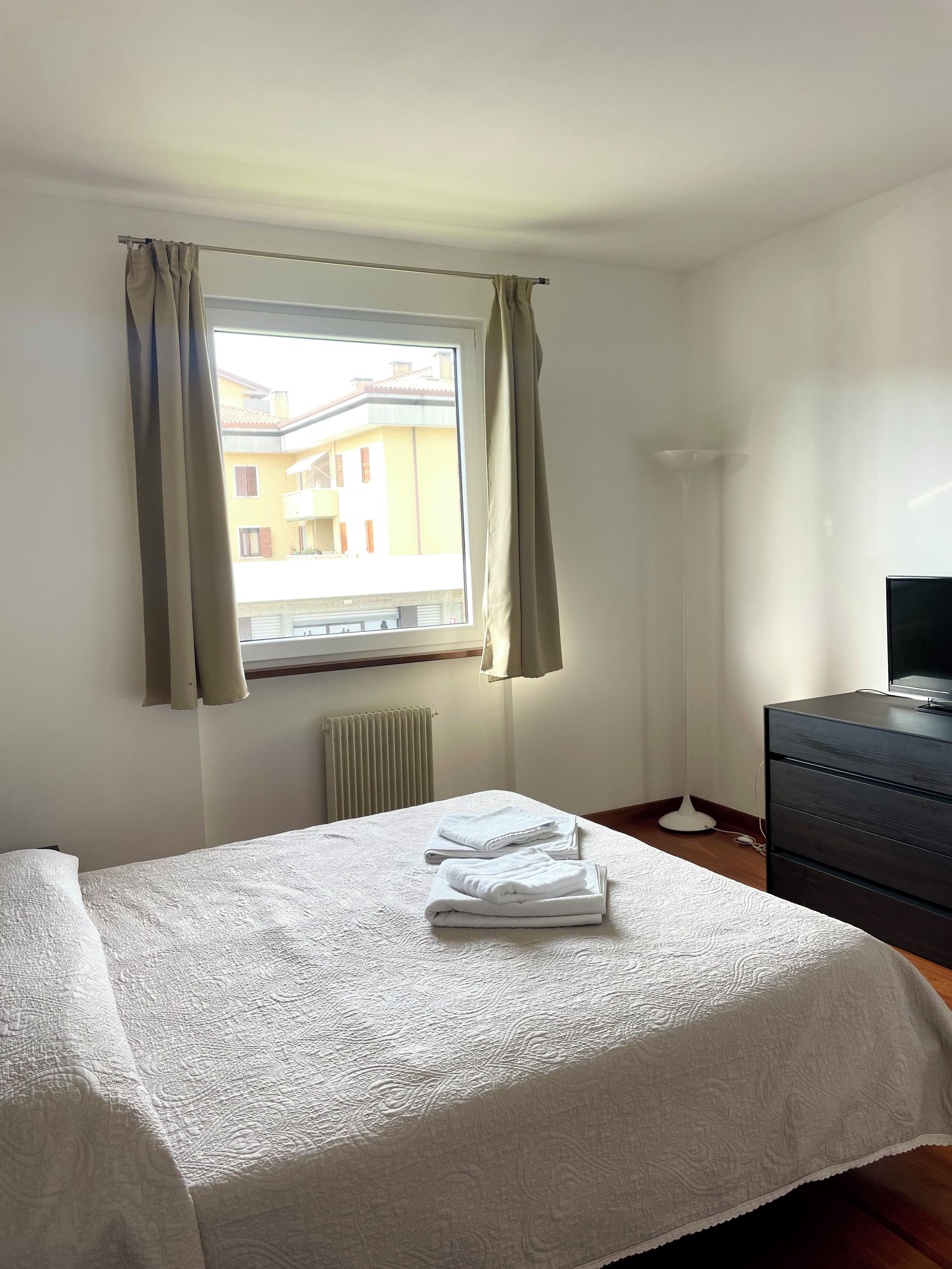 Quarto de hotel com cama, cômoda, radiador e janela com cortinas.