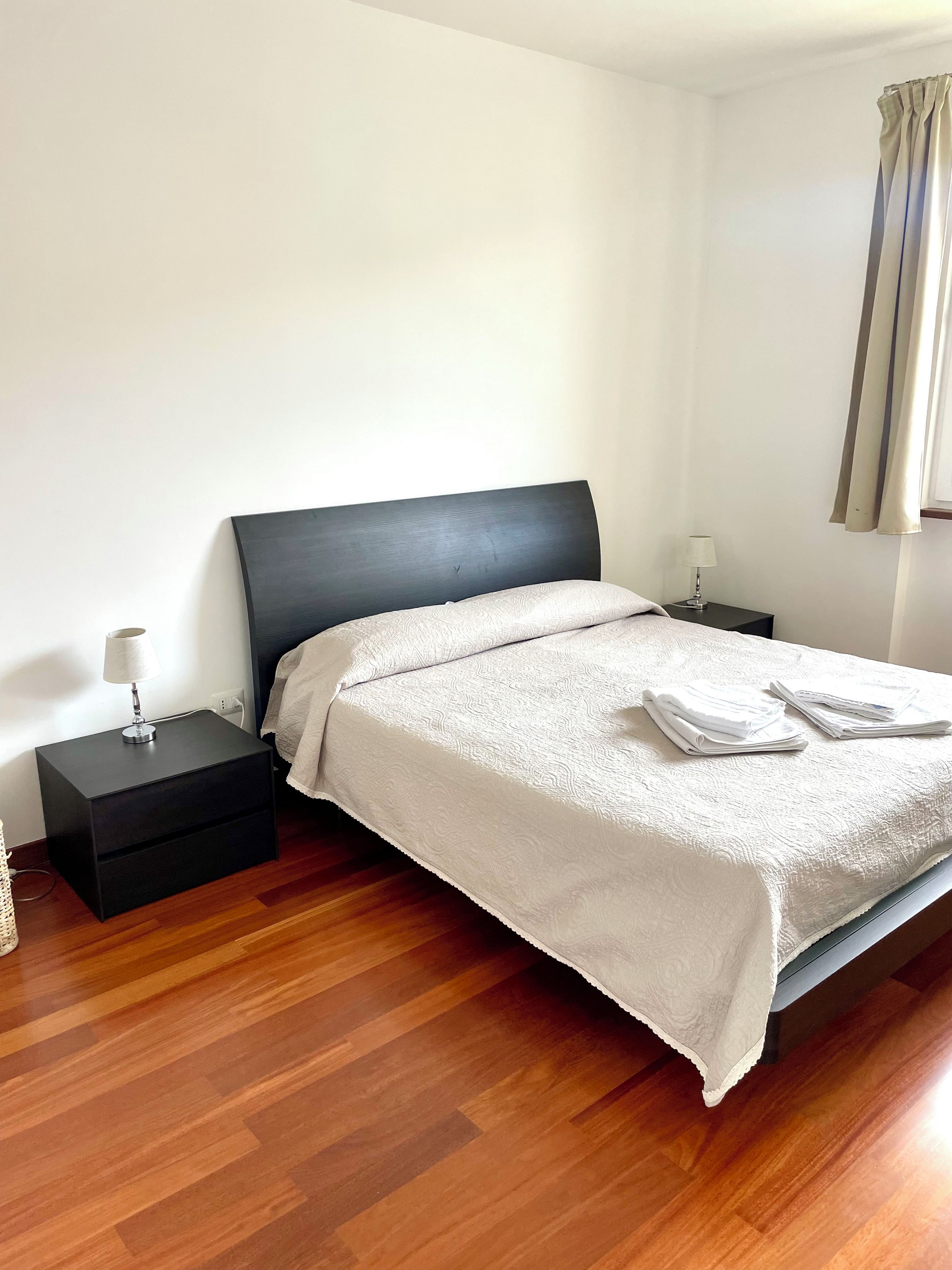A neatly made bed with a white bedspread and folded towels, a black nightstand with a lamp, and a wooden floor.
