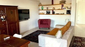 A living room with a sofa, a red armchair, a dining table with chairs, a TV, and shelves with various items. - HOLIDAY APARTMENT IN THE HEART OF VALPOLICELLA (San Pietro In Cariano)