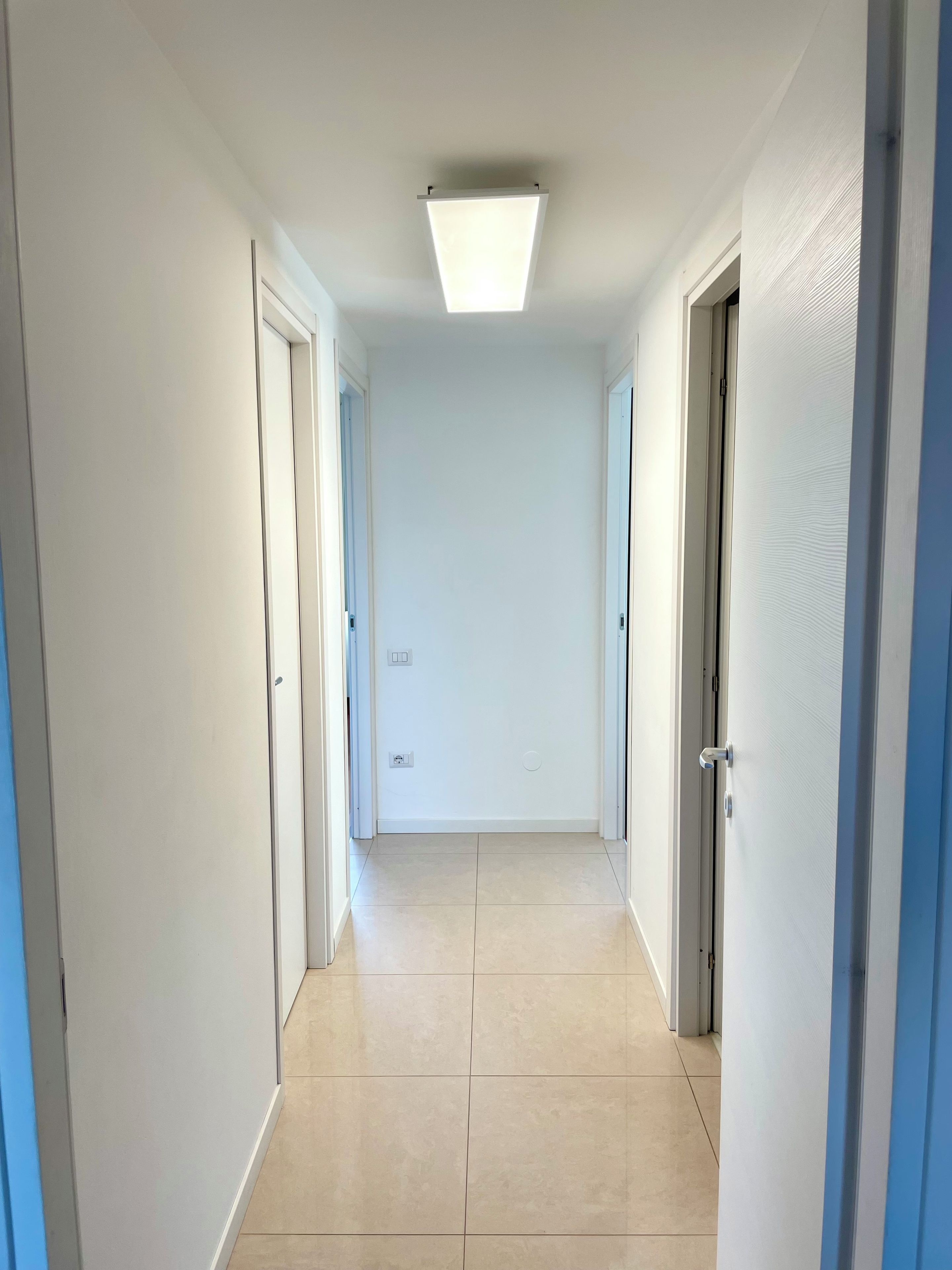 Un couloir aux murs blancs, doté d’un sol carrelé et d’un luminaire au plafond.