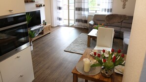 A modern living room with a sofa, a dining table set with a vase of red tulips, and a television. - Apartment "Am See" 2 (Rhauderfehn)