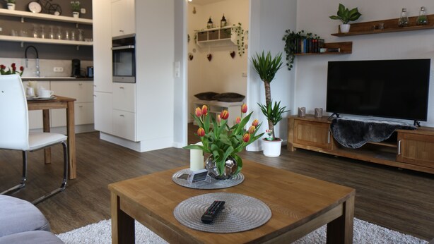 A modern living room with a wooden coffee table, a TV, and a kitchen area in the background. - Apartment "Am See" 2 (Rhauderfehn)