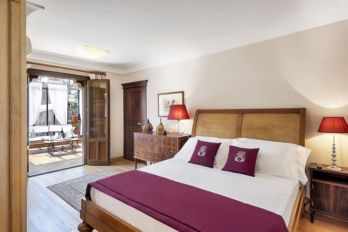Ein Schlafzimmer mit einem großen Bett, zwei Nachttischen, einem kleinen Tisch, einem Holzschrank, einer Tür zum Balkon und einem gerahmten Bild an der Wand.