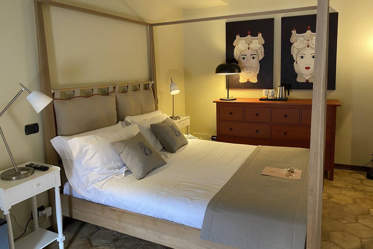 Ein Himmelbett mit Baldachin, ein Nachttisch mit Lampe, ein Beistelltisch, eine Holzkommode und zwei gerahmte Kunstwerke an der Wand.