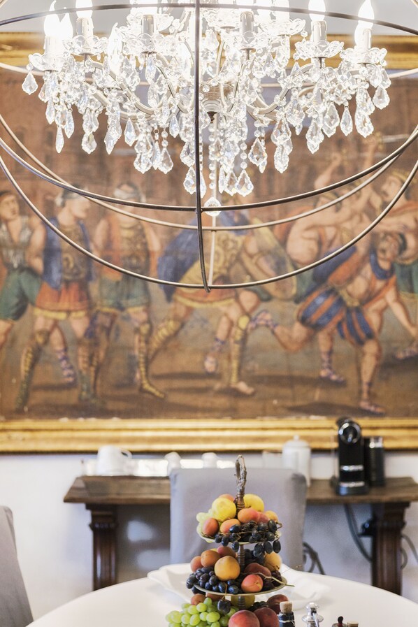 A chandelier with multiple crystal elements, a table with a fruit centerpiece, and a classical painting in the background. - Relais 147 (Taormina)
