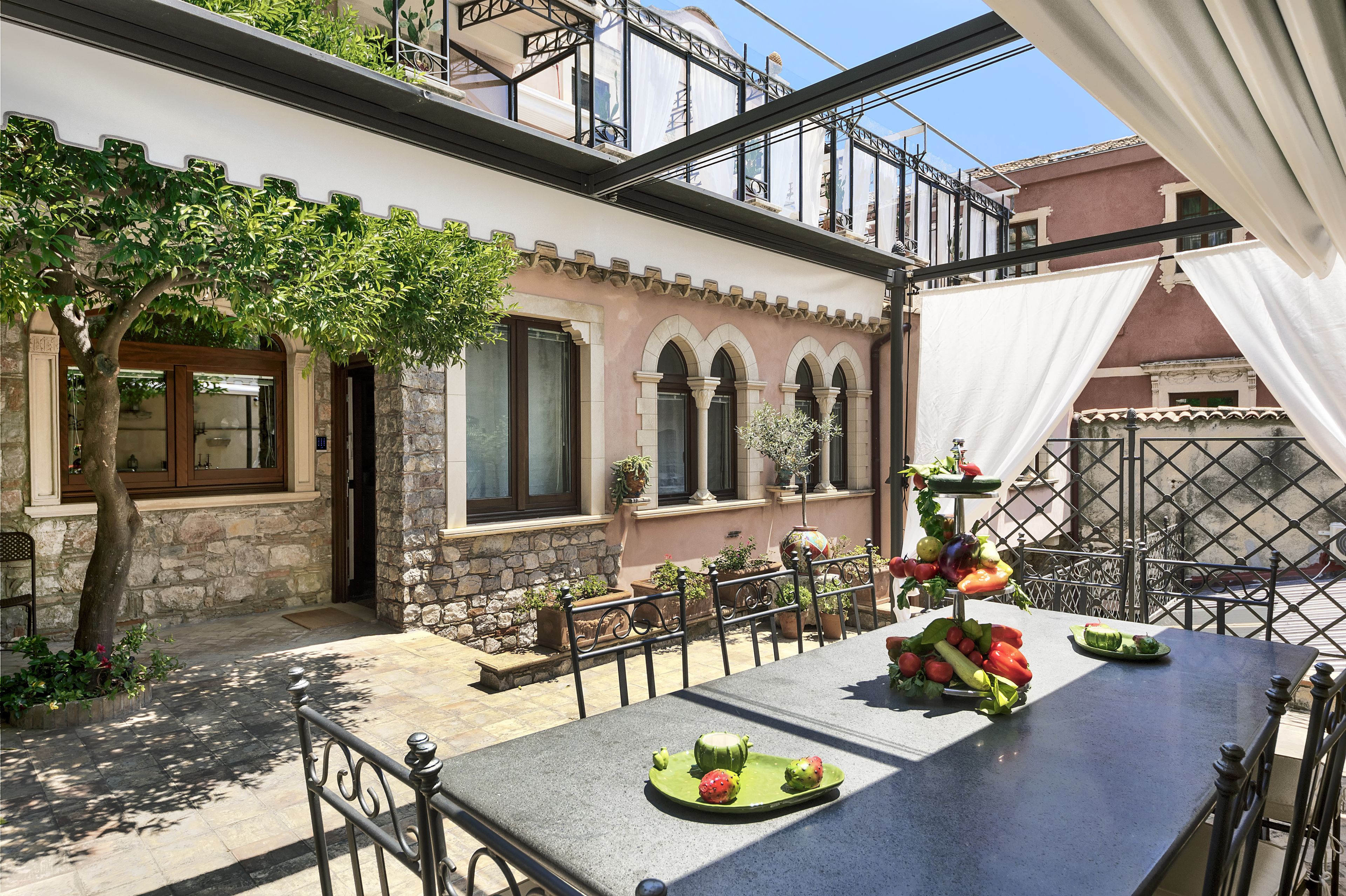 Un patio con una mesa dispuesta con fruta, un árbol y un edificio de piedra con arcos.
