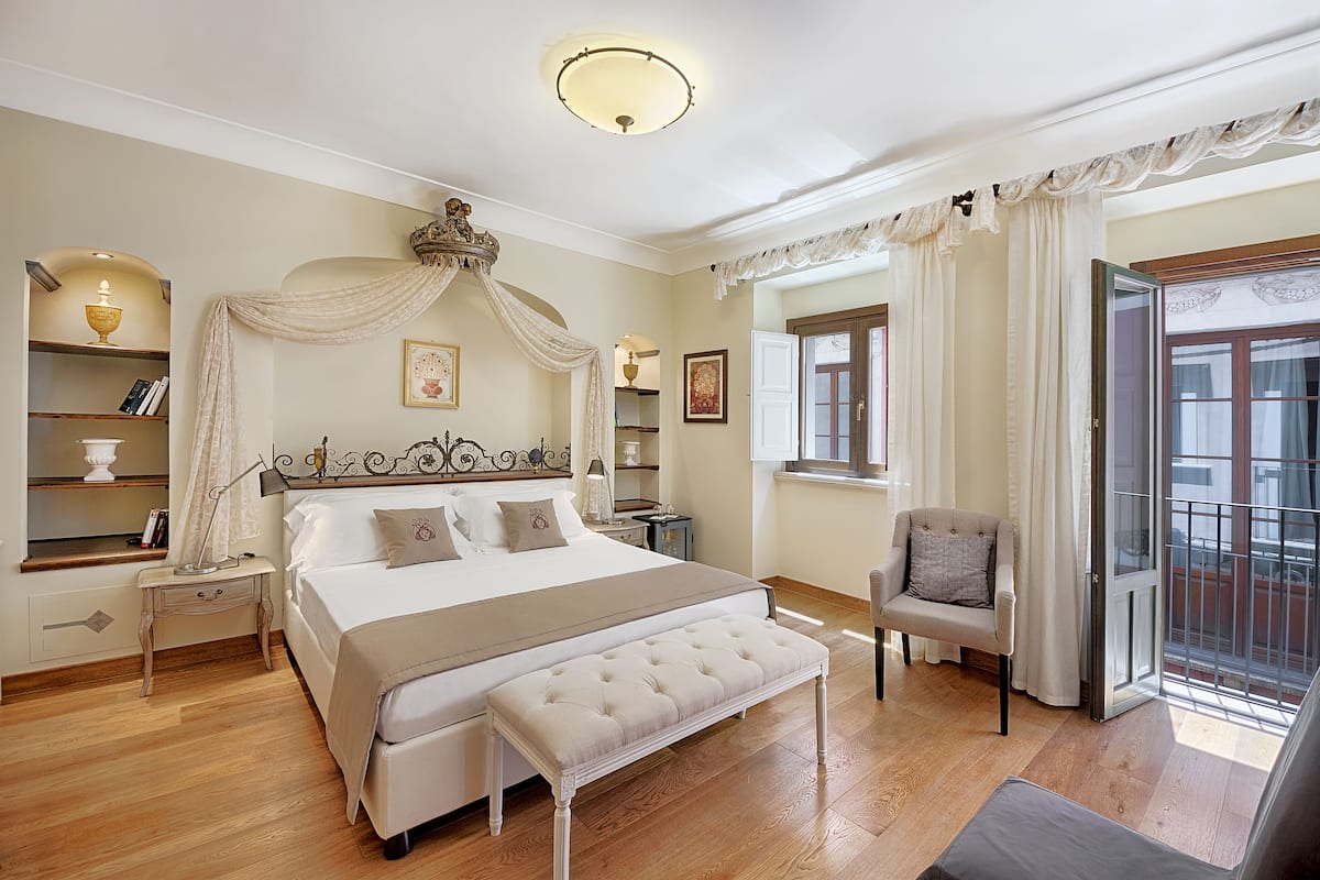 Ein geräumiges Schlafzimmer mit einem großen Bett, einem Nachttisch, einer Bank, einem Stuhl und einem Balkon mit Vorhängen.