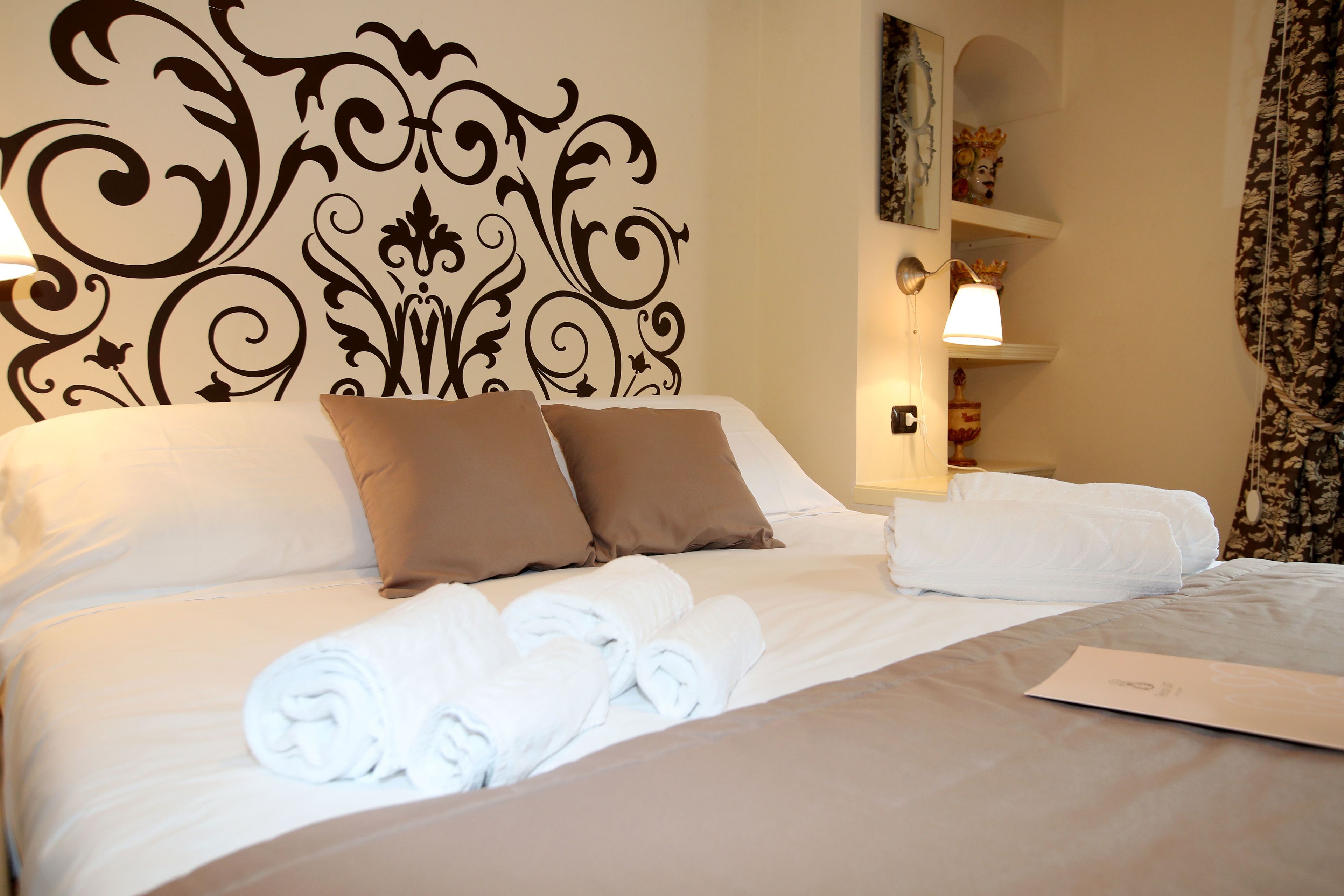 A neatly made bed with white and brown linens, a decorative headboard, and a wall-mounted shelf with a lamp.