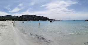 A beach with clear water, boats, and a hilly landscape in the background.