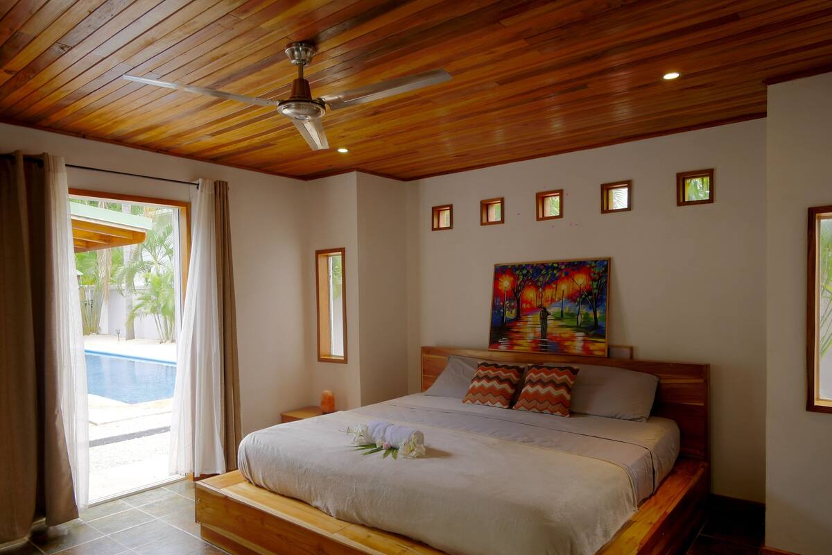 A bedroom with a wooden ceiling, a bed with a white bedspread, a painting above the bed, and a view of a swimming pool through a door.