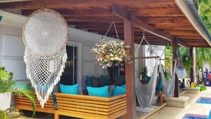 Un espacio al aire libre techado con un banco de madera, decoraciones colgantes de macramé y una piscina.