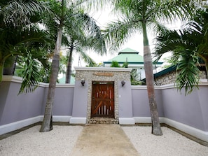 Une entrée fermée par un portail, avec un mur en pierre et un portail en bois, le tout entouré de palmiers et, au fond, d’un bâtiment doté d’un toit végétalisé.