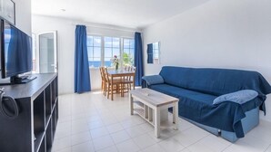 A modern living room with a blue sofa, a wooden coffee table, a TV on a black stand, and a dining area with a table and chairs. - Vista Mar Apartamentos (Tías)