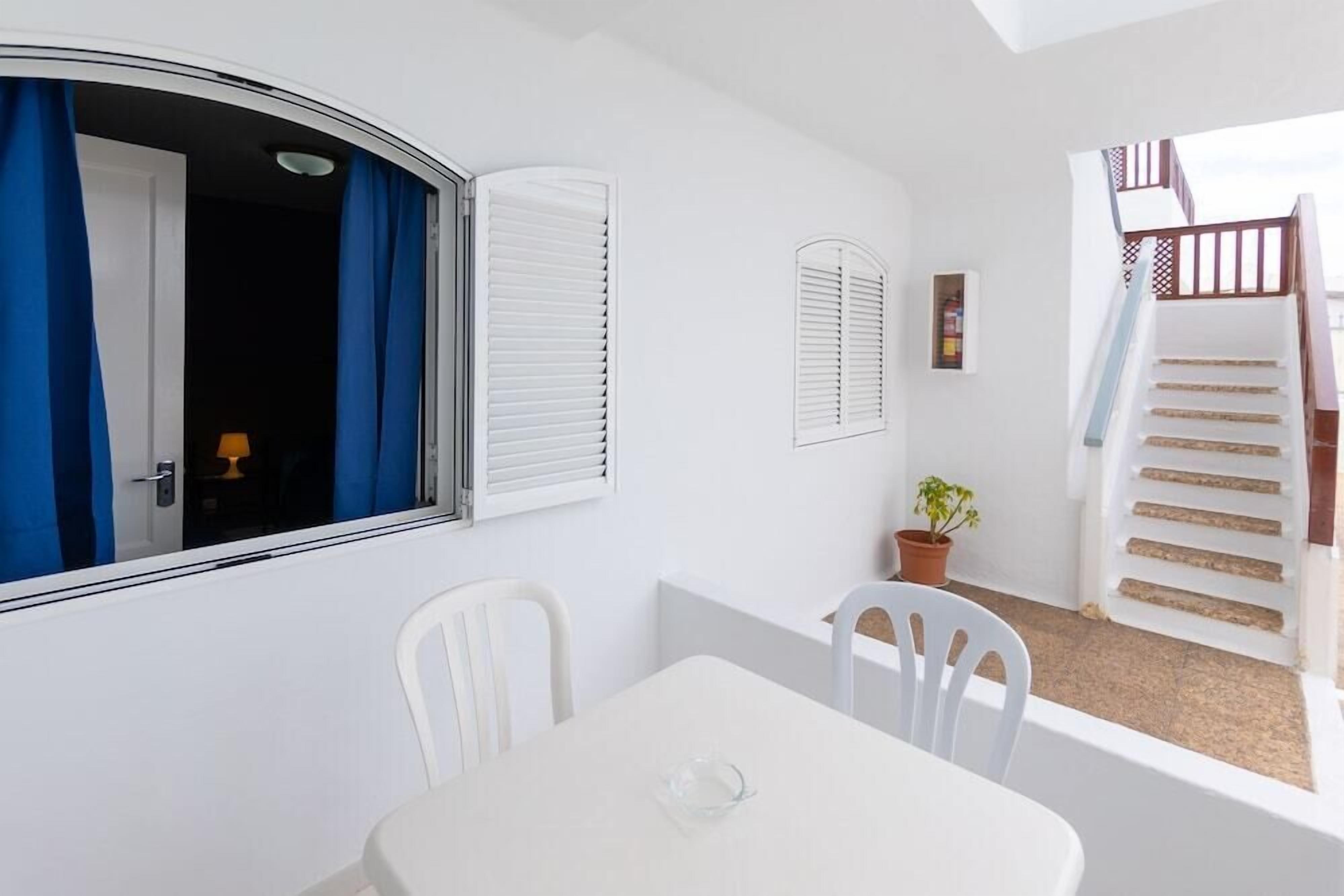 A white interior with a table and chairs, a window with blue curtains, and a staircase with white railings.