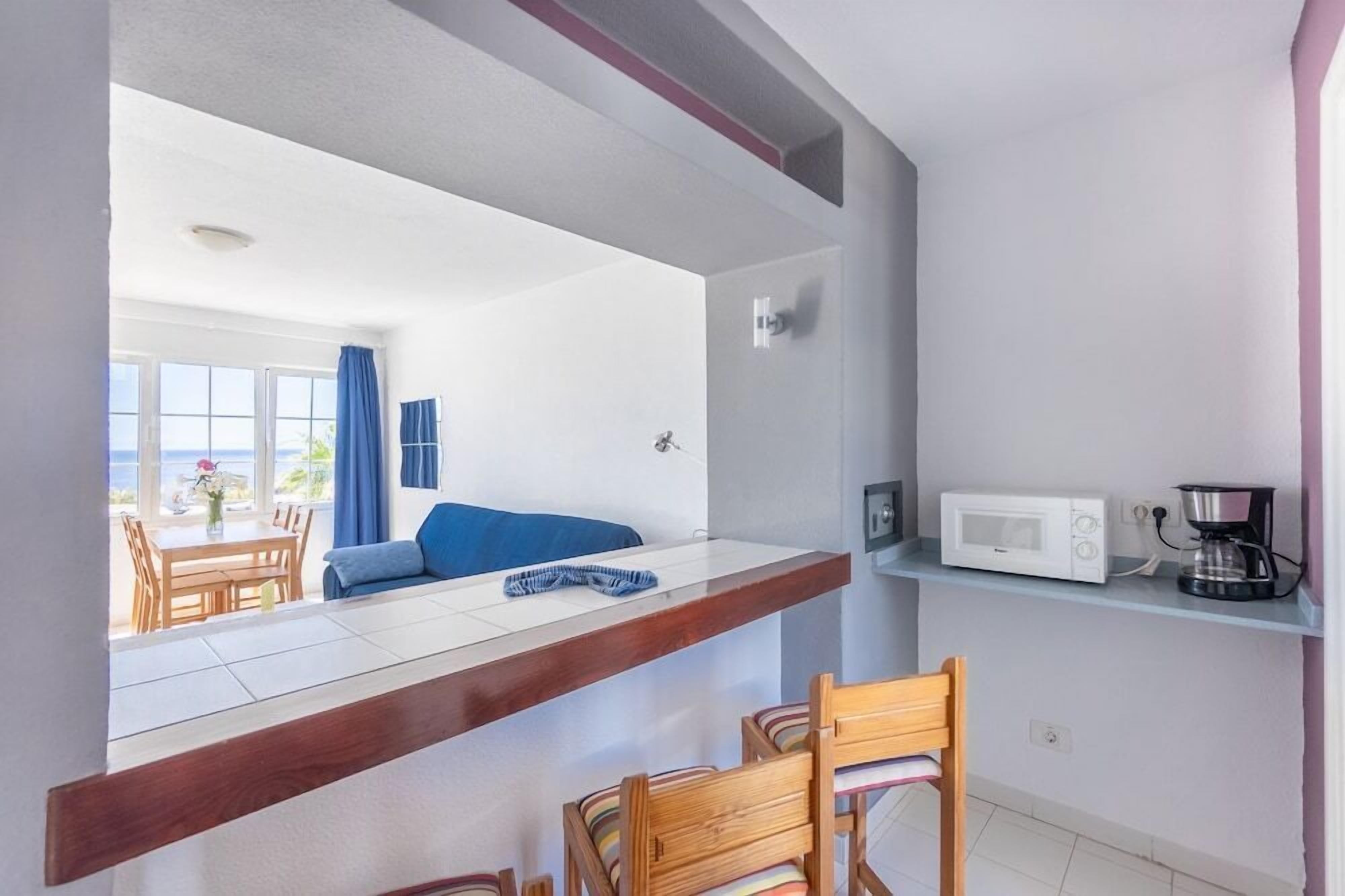 A compact kitchen with a microwave, coffee maker, and a dining area with a blue sofa and wooden chairs.