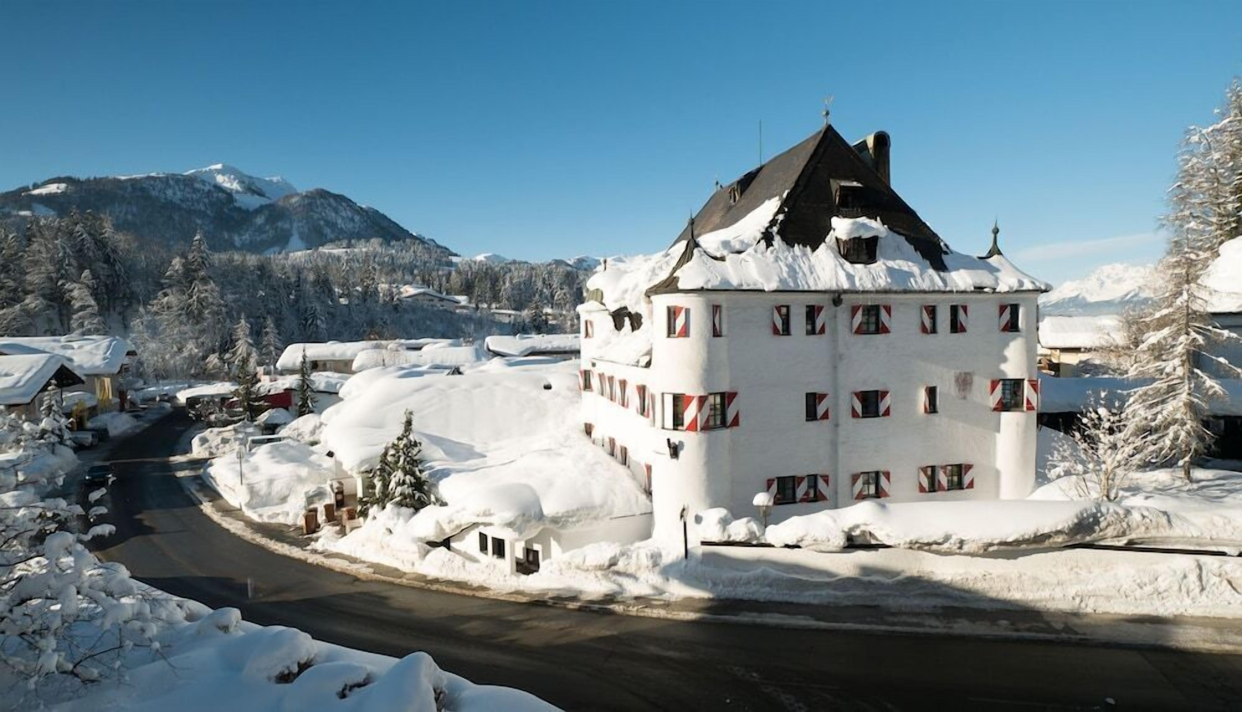 A white building with multiple windows and a snow-covered roof, surrounded by snow-covered trees and a mountain in the background.